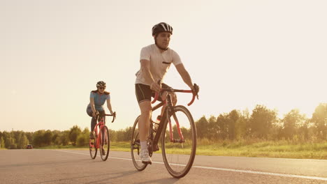 Dos-Ciclistas,-Un-Hombre-Y-Una-Mujer,-Viajan-Por-La-Carretera-En-Bicicletas-De-Carretera-Con-Cascos-Y-Ropa-Deportiva-Al-Atardecer-En-Cámara-Lenta